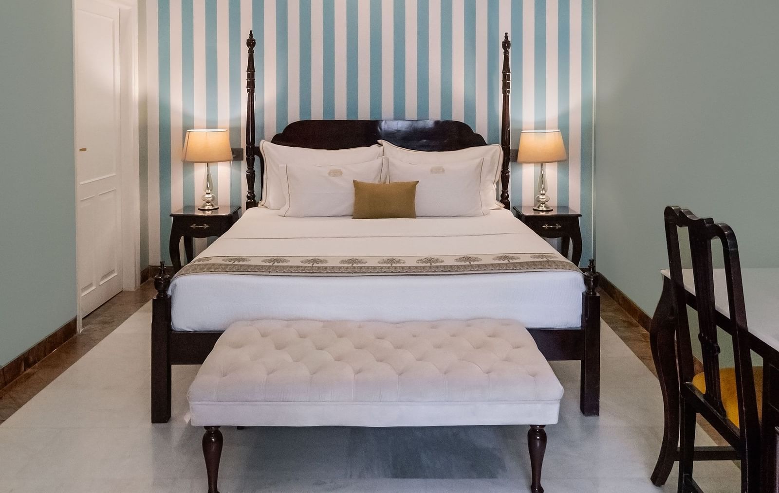 A narrow bedroom with a four-poster bed, vertical striped wallpaper, pale green walls, and a white floor, with a bench and chairs on either side | Dileep  Kothi, Jaipur